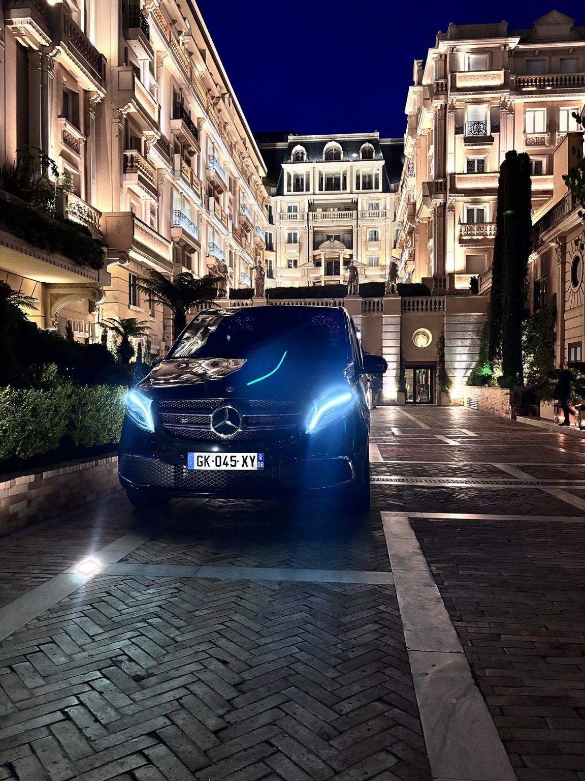 Chauffeur AMB Élite accueillant un client devant un palace de la Côte d'Azur — Mercedes Classe V de nuit